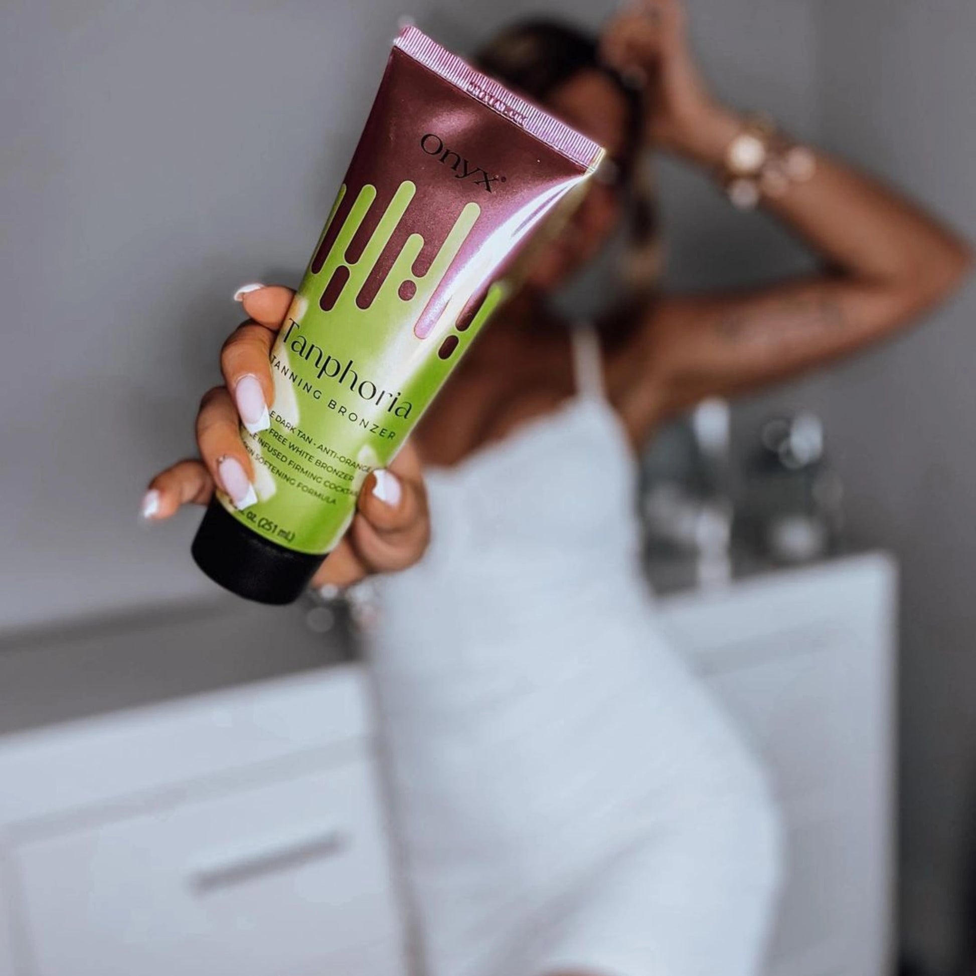 A woman in a white dress holding Onyx Tanphoria Bronzer Lotion in focus, with a blurred background.
