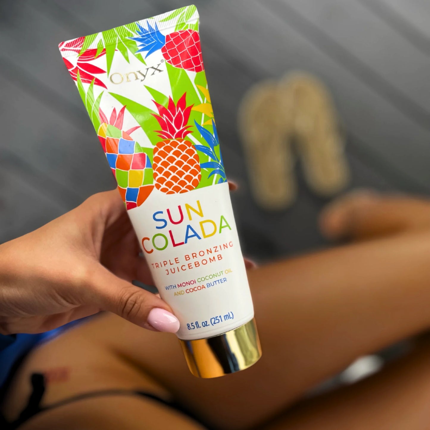 Hand holding Onyx Suncolada Triple Bronzing Juicebomb tanning lotion with tanned legs in the background