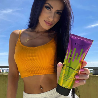 A model in a yellow crop top holding Onyx Tanphoria Bronzer Lotion under a bright blue sky.